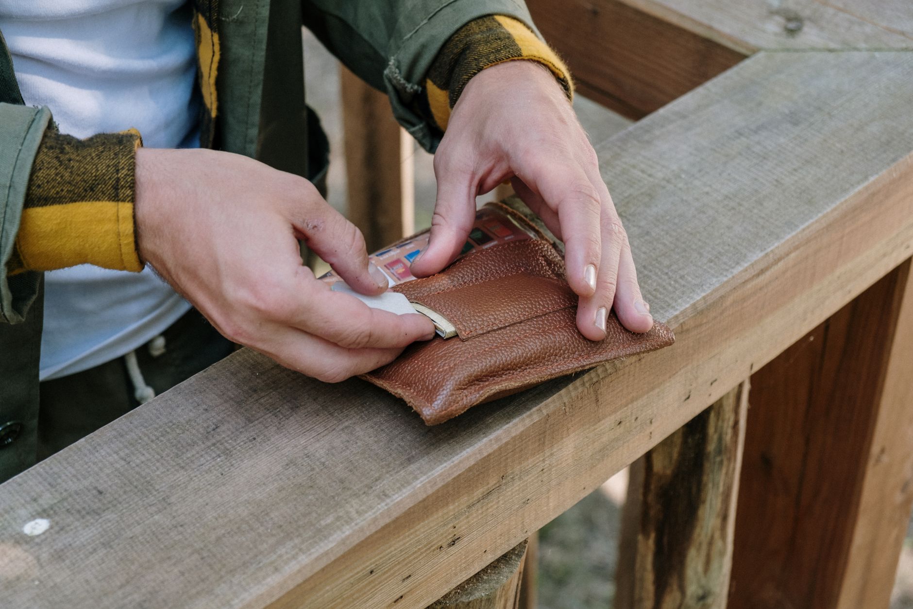 A man handling a leather wallet on a wooden railing, showcasing outdoor rustic style.