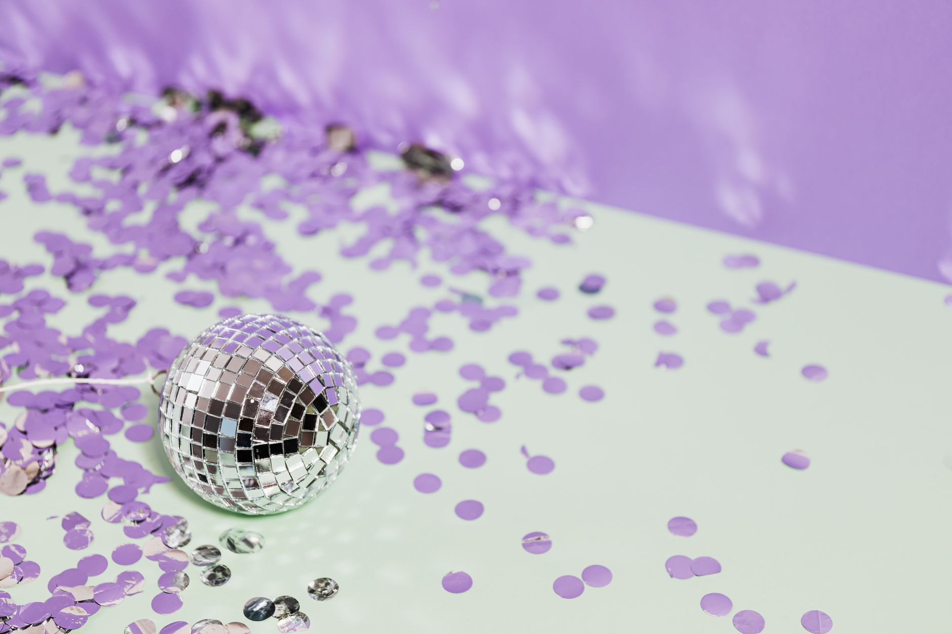 A shiny disco ball surrounded by purple confetti on a pastel background, evoking a festive mood.