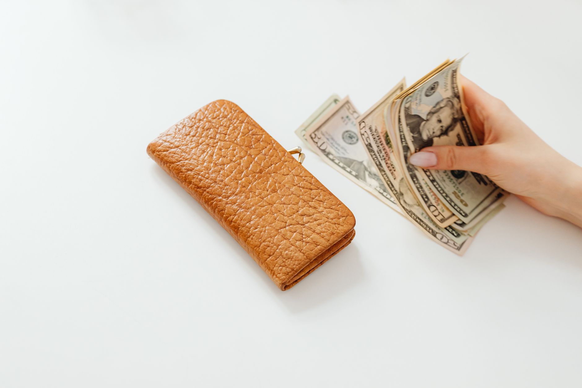 A hand counting American dollar bills next to a brown leather wallet on a white surface.
